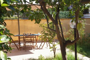 Eine Terrasse im Freien mit Tisch und Stühlen, umgeben von Grünpflanzen und einer gelben Wand.
