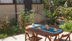 Un patio esterno con un tavolo rotondo in legno, sedie e una tovaglia. Sul tavolo ci sono due tazze bianche e un teiera.