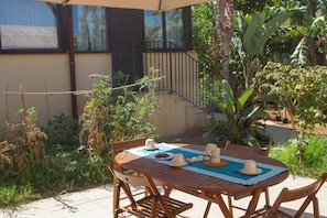 Eine Terrasse im Freien mit einem runden Holztisch, Stühlen und einer Tischdecke. Auf dem Tisch stehen zwei weiße Tassen und ein Teekessel.
