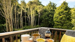 Ein Tisch draußen gedeckt mit verschiedenen Speisen und einer Flasche, umgeben von einer Holzterrasse und Bäumen im Hintergrund.