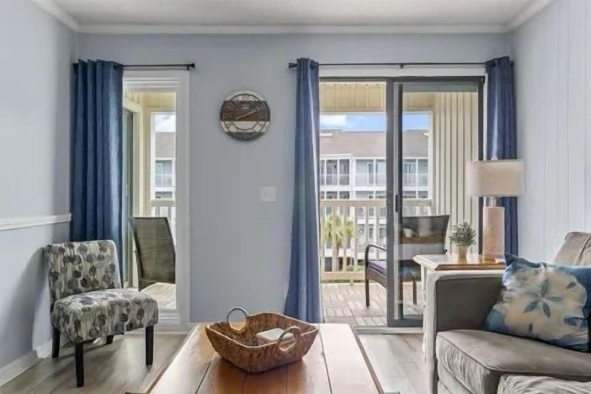 Una habitación con sofá, una silla, una mesa con una cesta y vistas a un espacio exterior por una puerta de cristal.