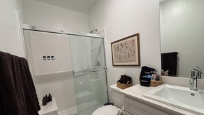 Et badeværelse med en glasafskærmet bruseniche, toilet, vask med spejl og et indrammet planteudsnit på væggen.