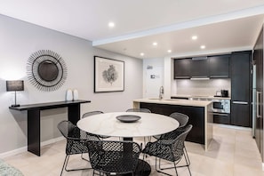 A modern kitchen with dark cabinetry, a central island, and a dining area with a round table and chairs. - Temple Escape 3 Bed Apartment with Private Pool (Craiglie)
