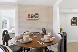 A dining area with a round table set for four, white bowls and plates, and a brick wall with a shelf above. - Modern Duplex Unit w/ Backyard, Near Transit (Seattle)