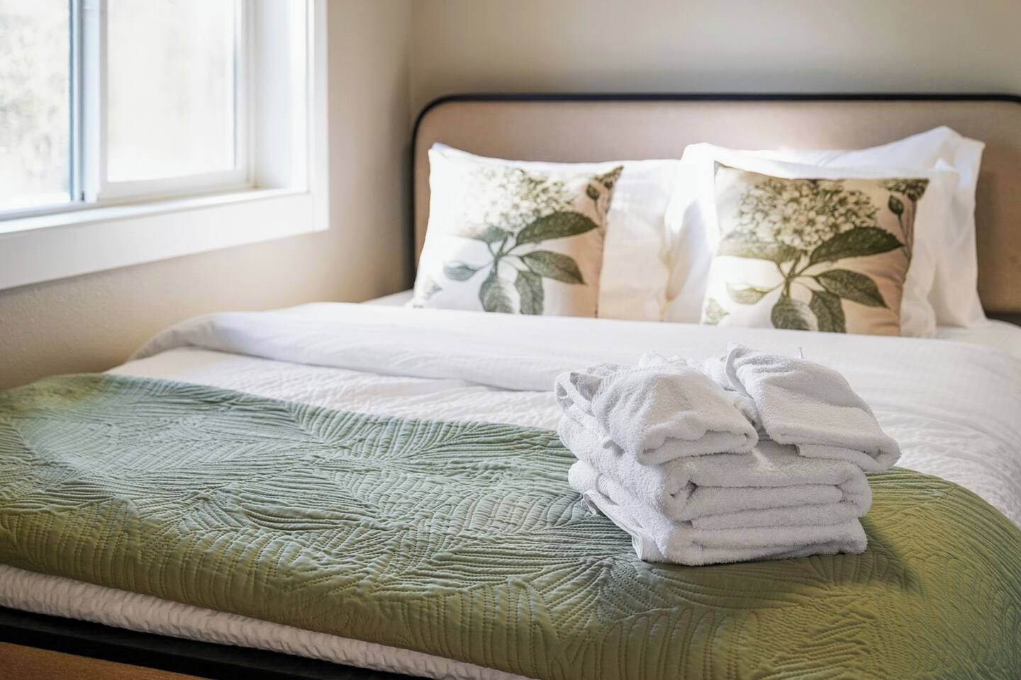 A neatly made bed with a green bedspread, two pillows with floral patterns, and a stack of white towels.