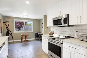 A modern kitchen with white cabinets, stainless steel appliances, and a window with a view of trees. - Modern Seattle Duplex, Balcony w/ Serene View (Seattle)