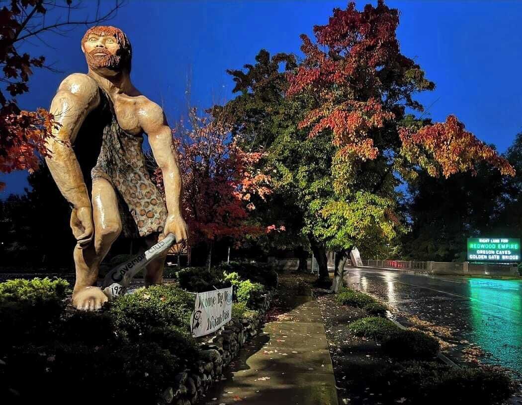 Una escultura de una figura musculosa con un cartel que dice 'Caveman Club' en un parque, con árboles y un sendero.