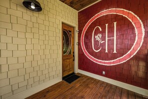 Un corridoio con una porta in legno, un muro rosso con uno stemma bianco e un lampadario sopra la porta.