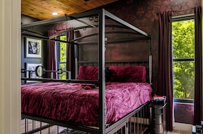 Ein Etagenbett mit Metallgestell und roter Bettwäsche, ein kleiner Nachttisch und ein Fenster mit Vorhängen.