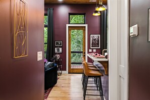 A modern interior with a wooden floor, a dining area with bar stools, a black sofa, and a large window with a view of greenery. - Adults-Only Couples Retreat · Private Hot Tub (Sevierville)