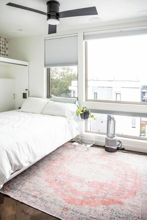 Quarto com cama, ventilador de teto, janela com persianas e uma pequena planta em um suporte.
