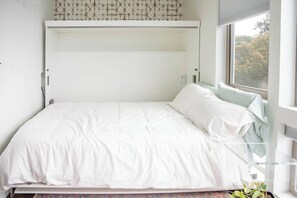 Ein weißes Bett mit schlichtem Kopfteil, ein Fenster mit Blick auf Bäume und eine gemusterte Wand über dem Bett.