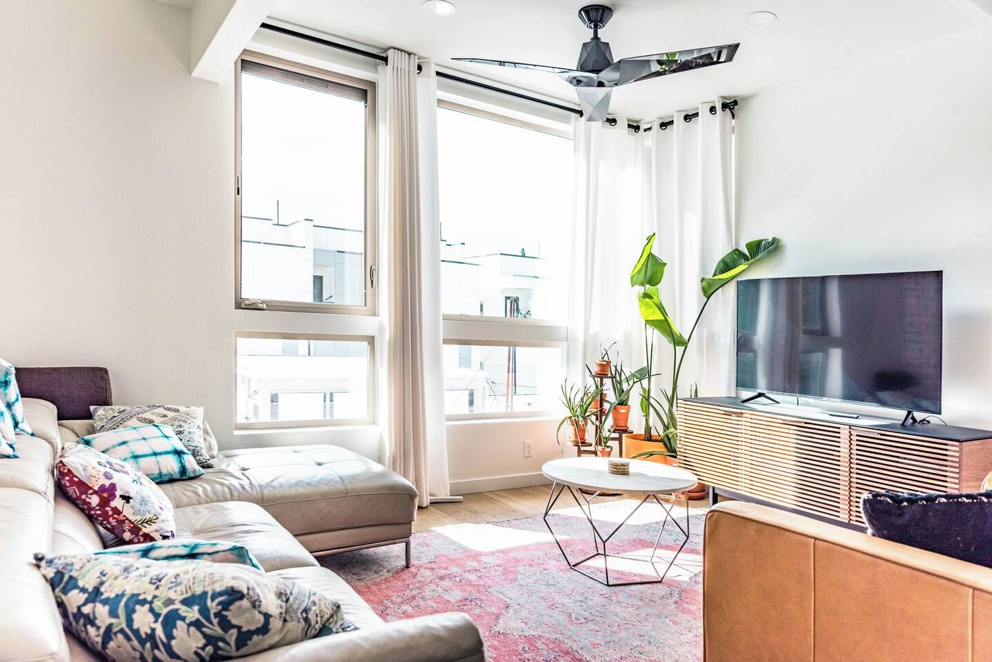 A modern living room with a sofa, a coffee table, and a TV. There are large windows and a ceiling fan.