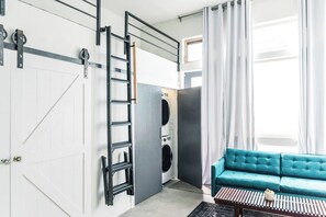 Ein kompakter Wohnbereich mit integrierter Waschmaschine und Trockner, einer Leiter zur Aufbewahrung, einem salbeengrünfarbenen Sofa und einem Couchtisch.