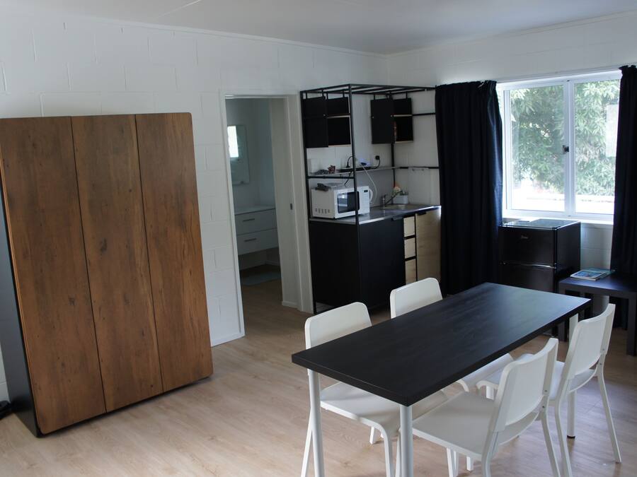 Cocina compacta con microondas, hervidor y armarios negros. Hay un comedor con una mesa negra y sillas blancas. Se aprecia un mueble de madera a la izquierda.