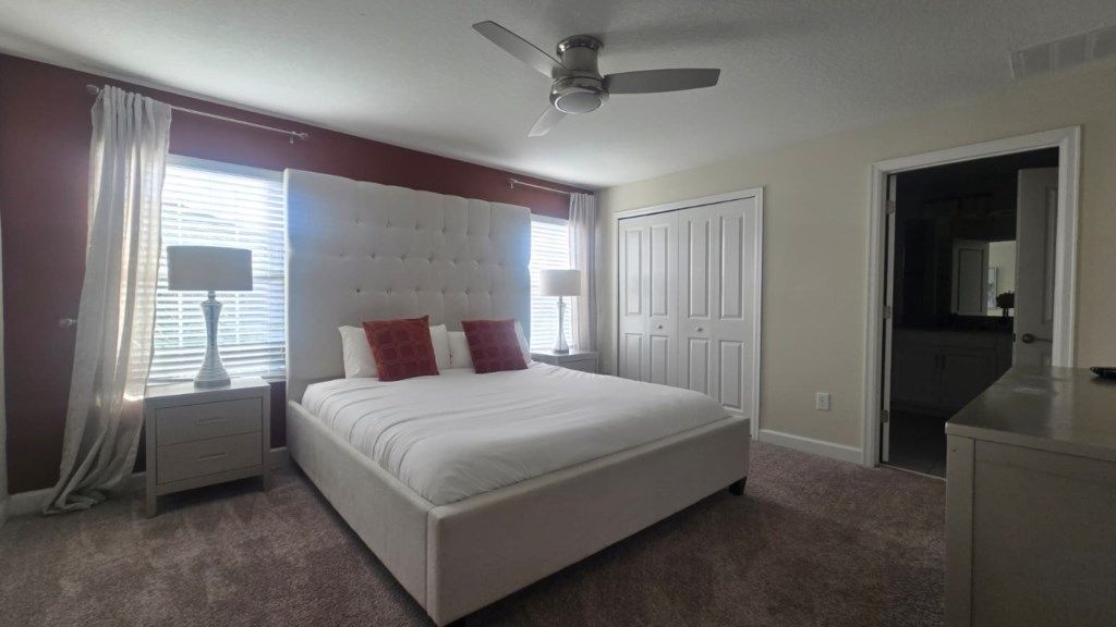 A bedroom with a large bed, a ceiling fan, a nightstand, a lamp, and a door leading to another room.