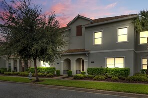 A two-story residential building with a symmetrical facade, a central entrance with an archway, and a well-maintained front yard with hedges and a tree. - Private pool, resorts, 9 minutes to Disney (Kissimmee)