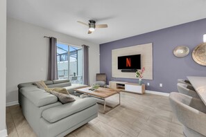 A modern living room with a grey sofa, a coffee table, a TV mounted on the wall, and a ceiling fan. - Private pool, resorts, 9 minutes to Disney (Kissimmee)