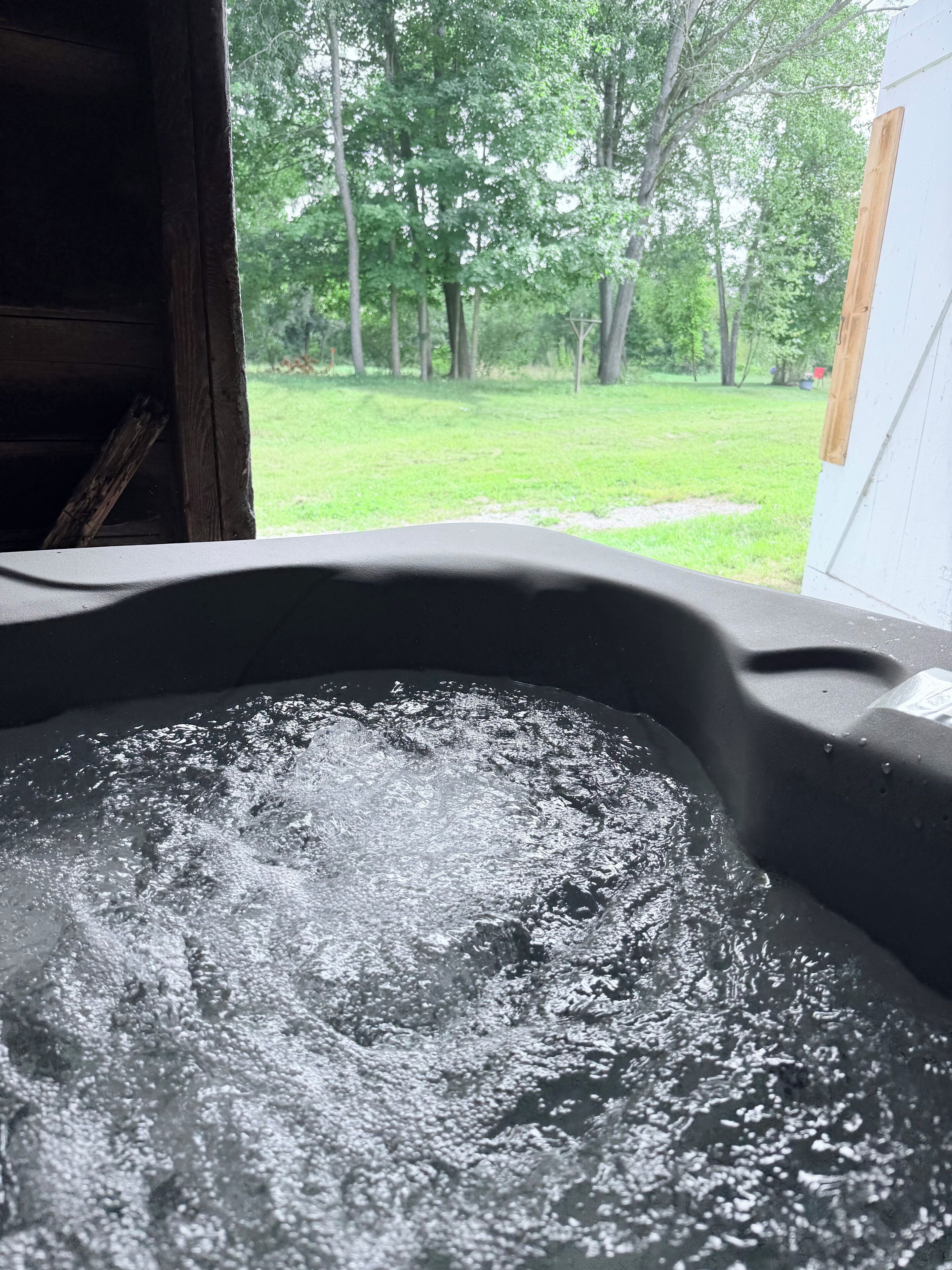 A hot tub with water bubbling, surrounded by a wooden deck and a grassy yard with trees.