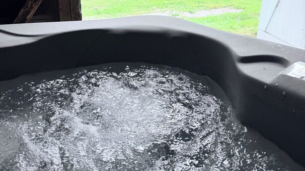 Un bain à remous où l’eau bouillonne, entouré d’une terrasse en bois et d’une pelouse verdoyante parsemée d’arbres.