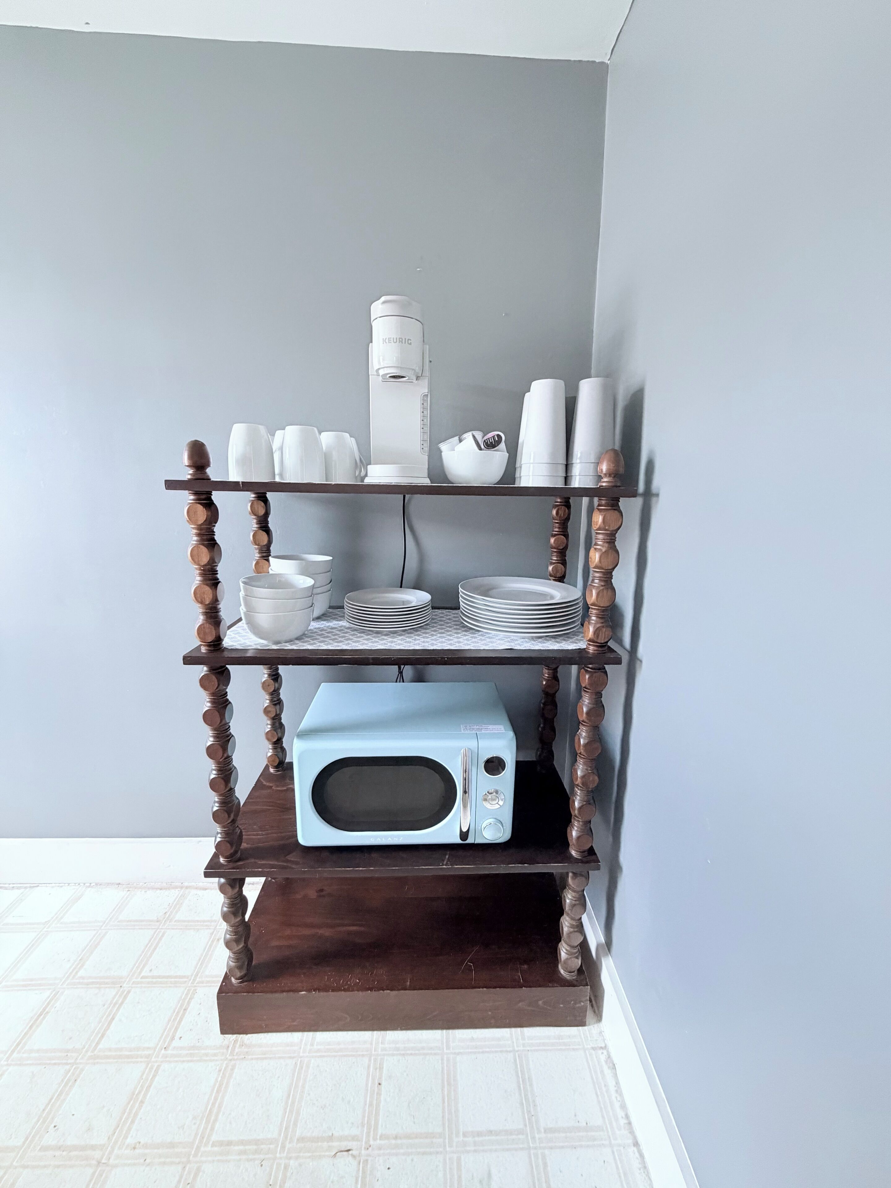 A kitchen area with a microwave on a wooden shelf, white cups, plates, and a coffee maker.