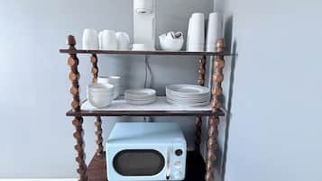 A kitchen area with a microwave on a wooden shelf, white cups, plates, and a coffee maker.