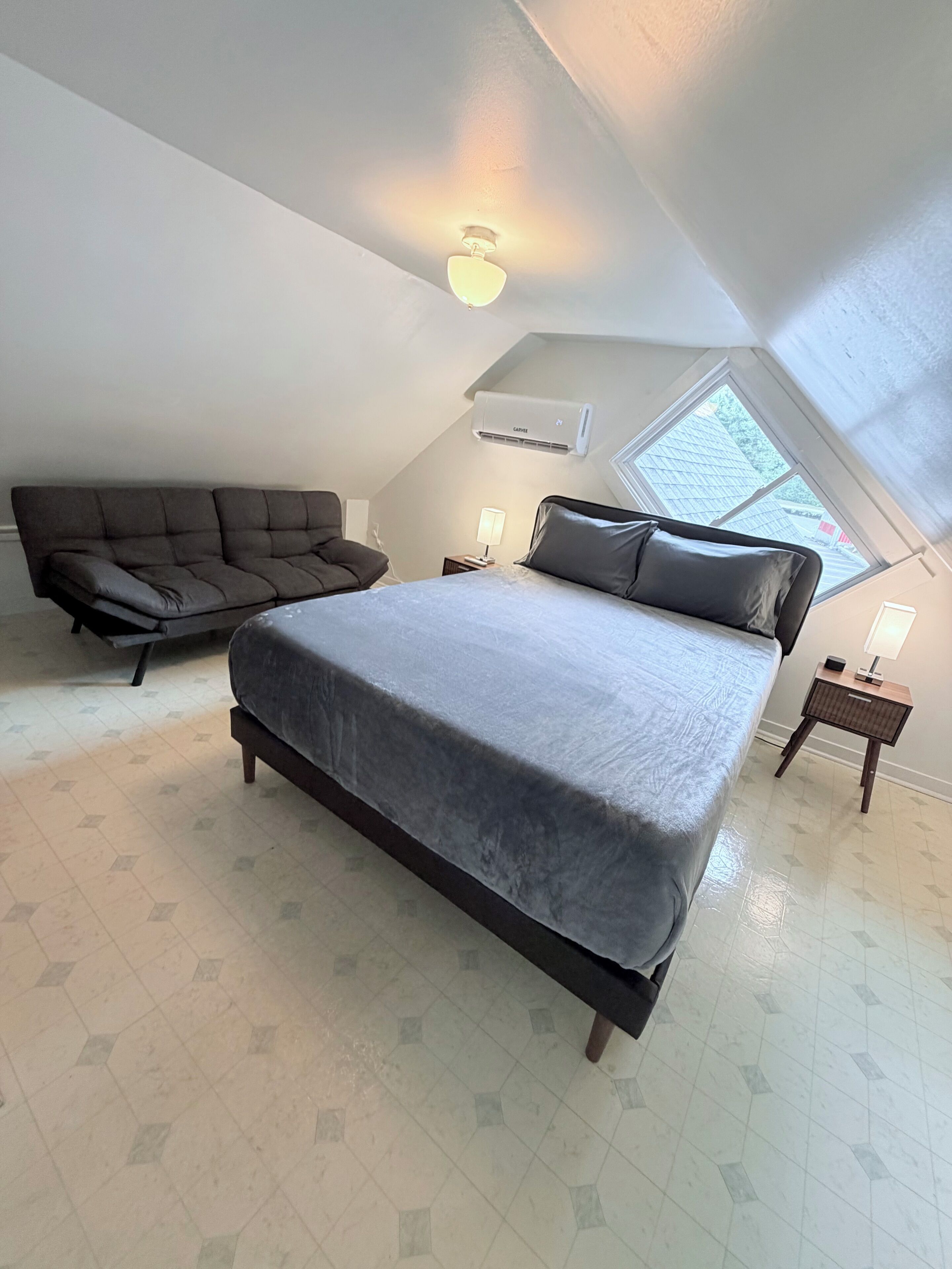 A bedroom with a large bed, a sofa, a nightstand with a lamp, and a skylight.