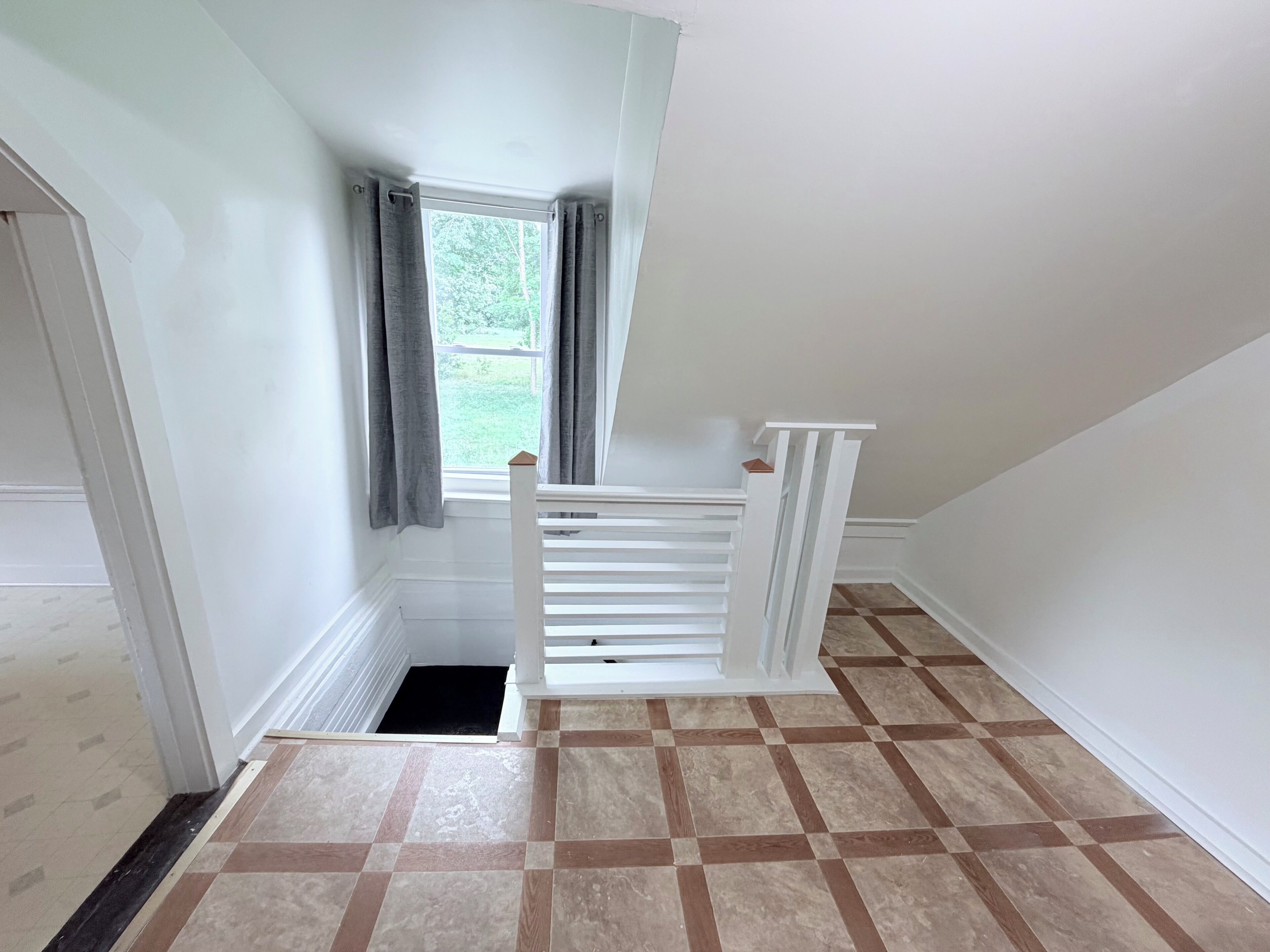 Une pièce avec un escalier blanc, une fenêtre avec des rideaux et un sol carrelé.