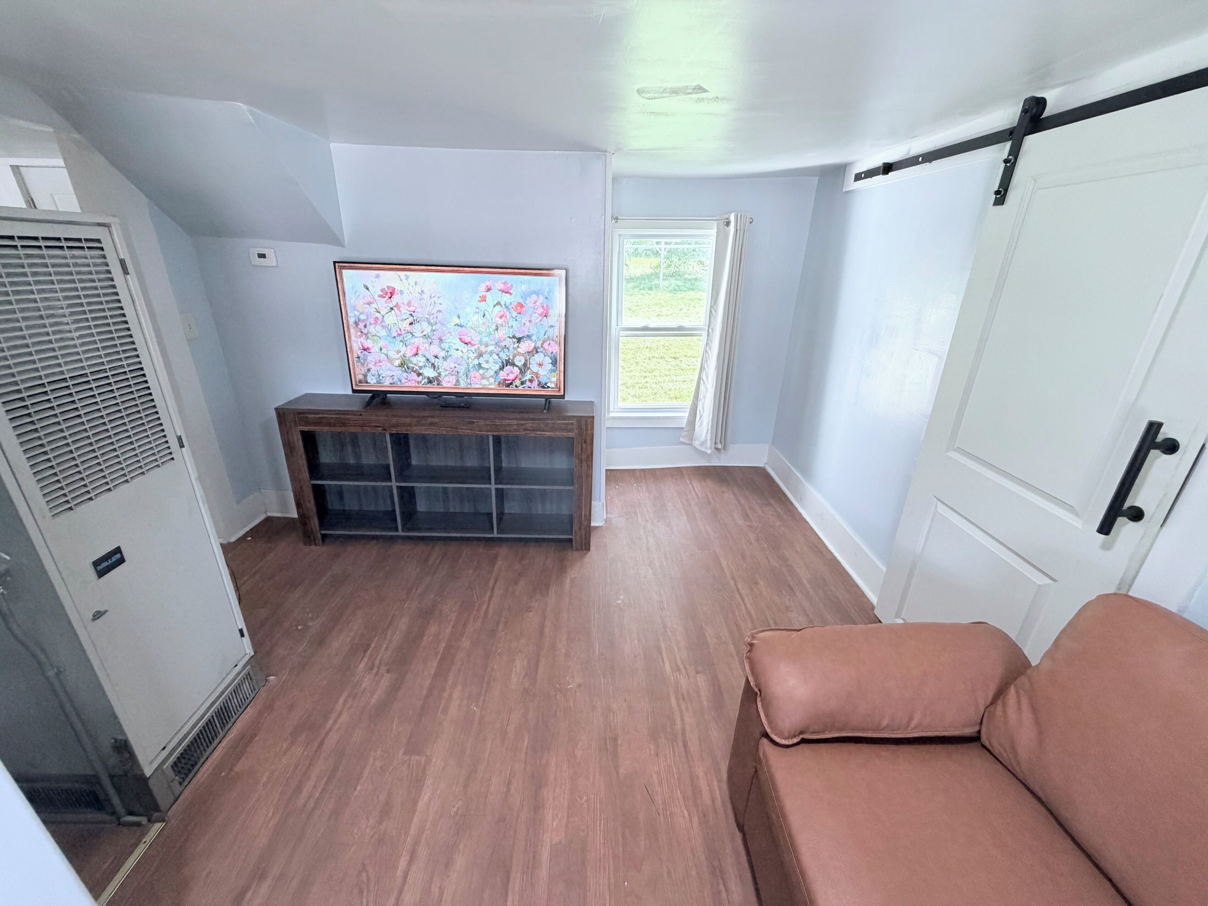 A compact room with a television on a wooden stand, a window with curtains, a door with a black handle, and a brown sofa.