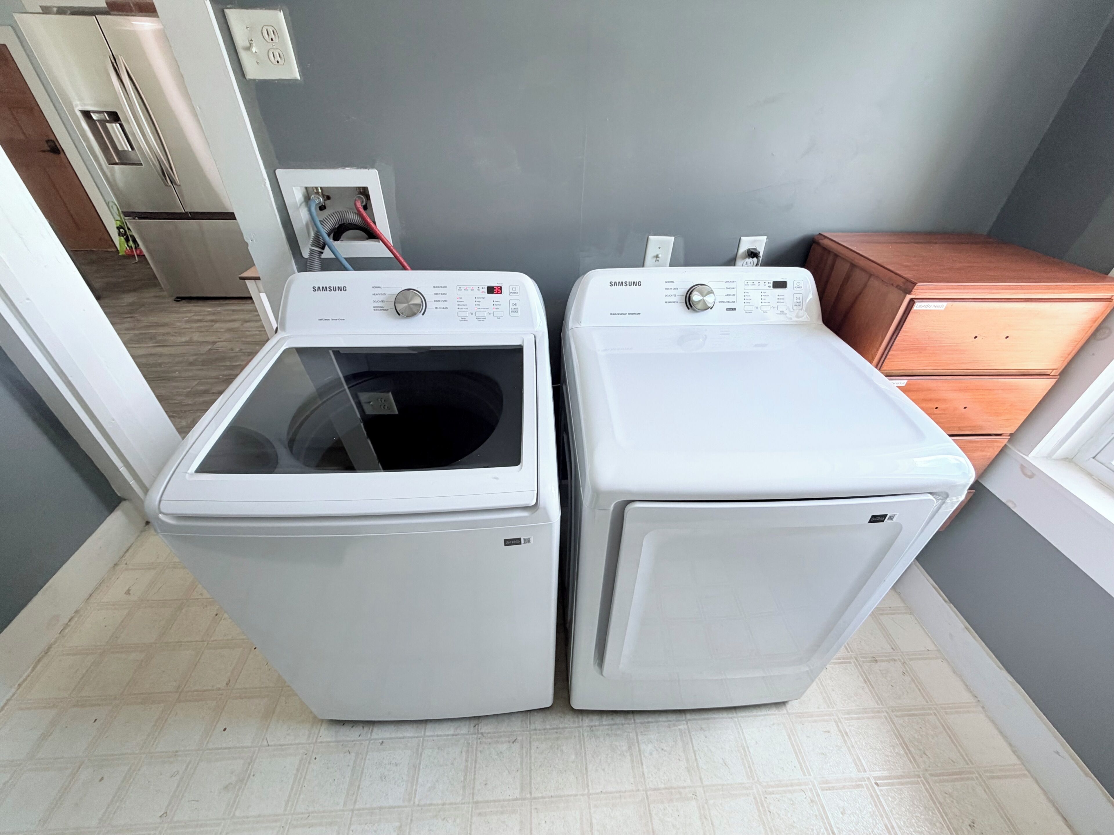 Une buanderie équipée de deux machines à laver Samsung blanches, d’un meuble en bois et d’un sol carrelé.