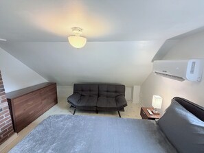 Une pièce comprenant un lit, un canapé sombre, une petite table avec une lampe, un ventilateur de plafond et un appareil de climatisation.