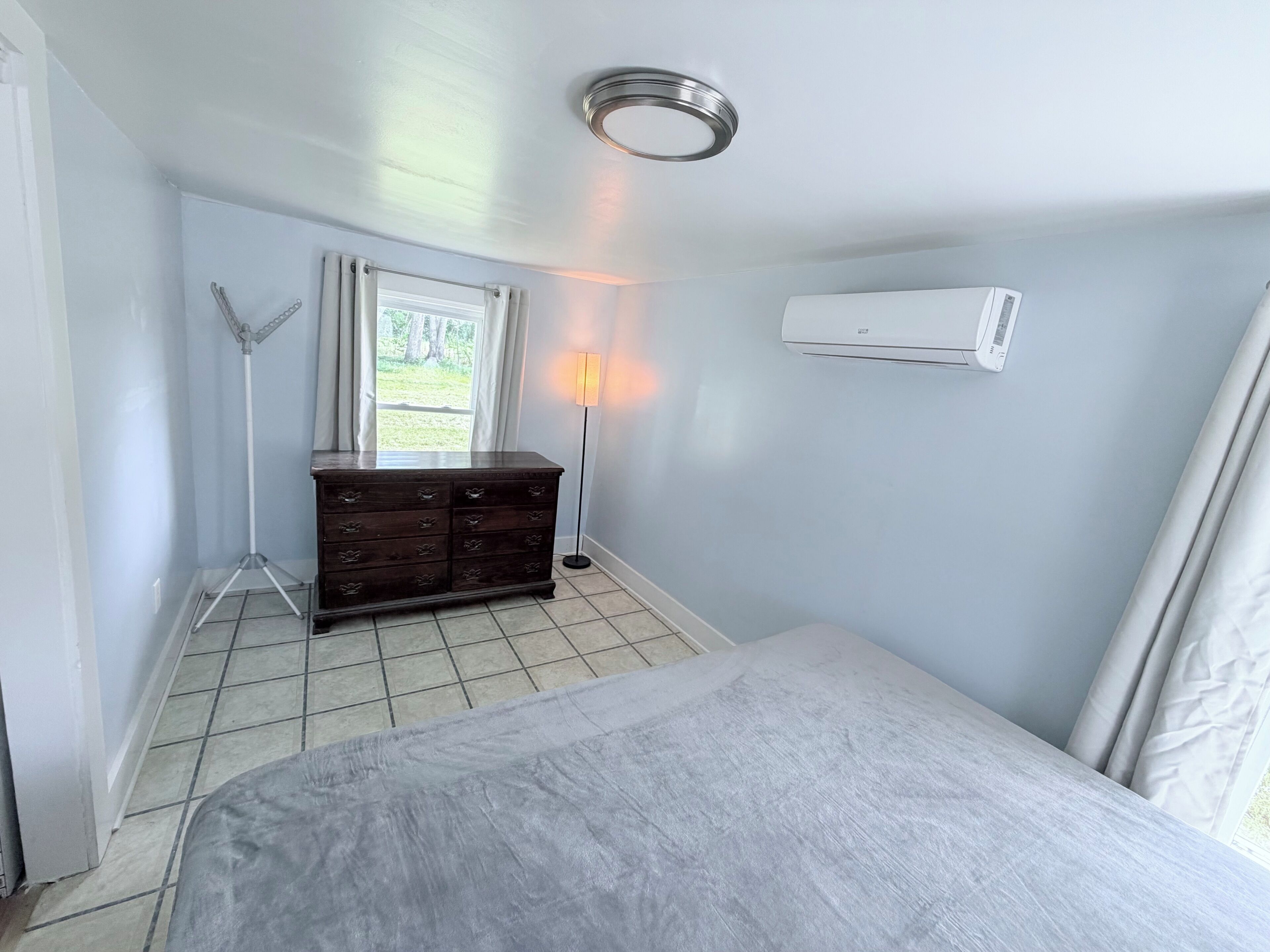 A room with a bed, a dresser, a lamp, and an air conditioning unit.