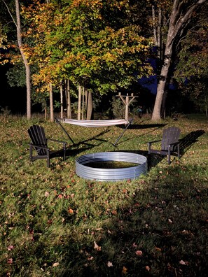 A hammock between two Adirondack chairs, a metal fire pit, and a grassy area with trees in the background.