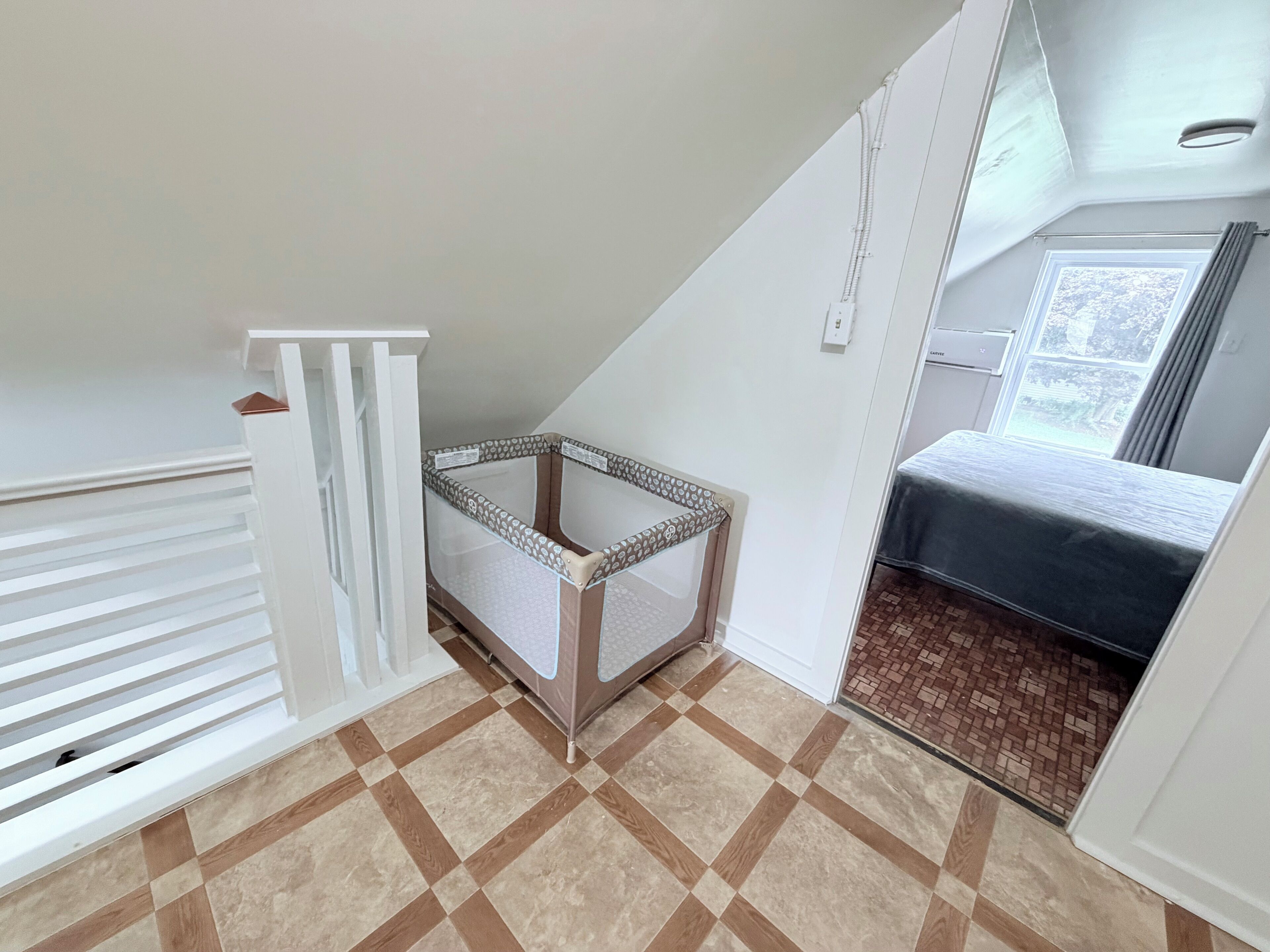 A room with a sloped ceiling, a crib, a bed, and a window with curtains.