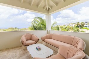 Ein Balkon mit zwei Sitzbereichen mit Polstern und einem kleinen Tisch, mit Blick auf eine Küstenlandschaft mit Palmen und Gebäuden.
