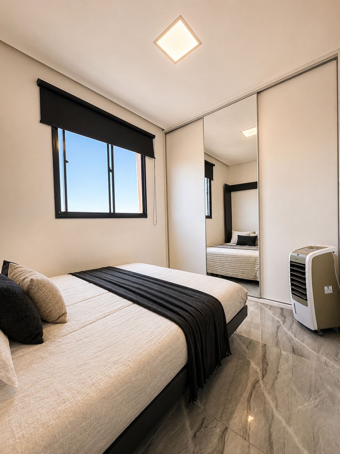 Quarto com cama, janela com cortina preta, espelho e ar condicionado portátil.