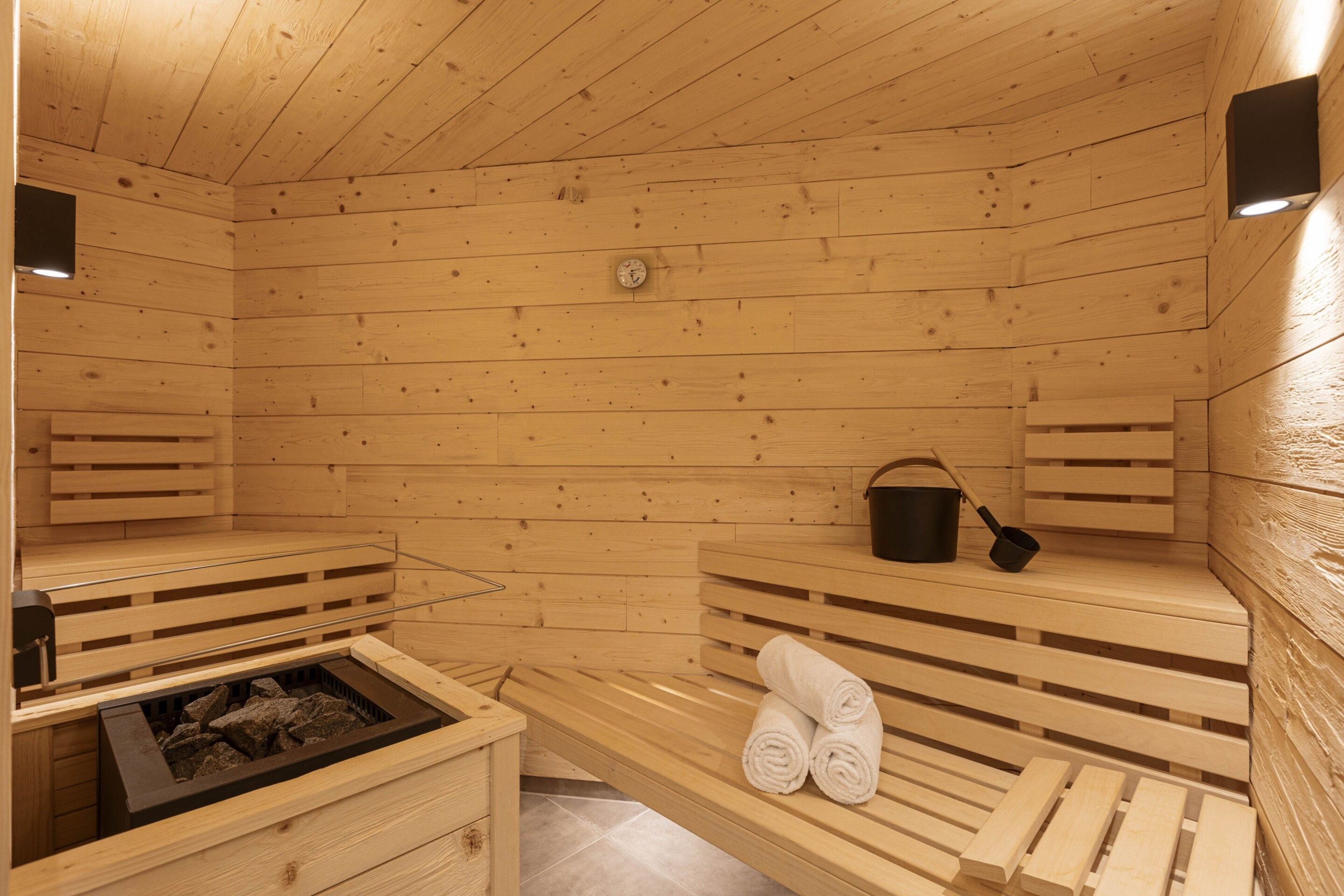 A sauna room with wooden benches, a bench with towels, a black bucket, and a heater.
