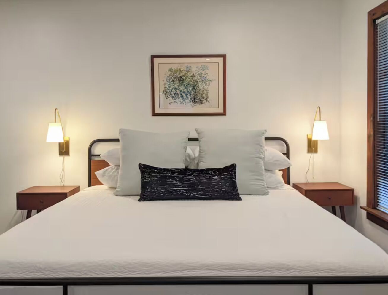 Een net opgemaakt bed met een witte dekbedovertrek, twee nachtkastjes met lampen, en een lijst met een kunstwerk aan de muur.
