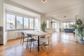 Un vaste espace de vie comprenant une grande table à manger, de nombreuses chaises et un canapé. On y trouve également plusieurs plantes en pot et, grâce aux grandes fenêtres, une vue sur les bâtiments environnants.