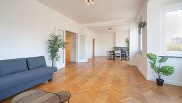 A spacious living area with a sofa, a dining table, and chairs, a kitchen in the background, and large windows.