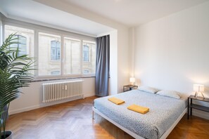 Une chambre avec un grand lit, deux tables de chevet avec des lampes, un radiateur, une fenêtre avec des rideaux et une plante en pot.