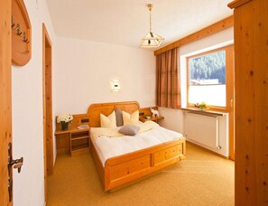A bedroom with a wooden bed, a nightstand, a lamp, a window with a view of trees, and a radiator. - Holiday Apartment With Wellness Area (Laengenfeld)