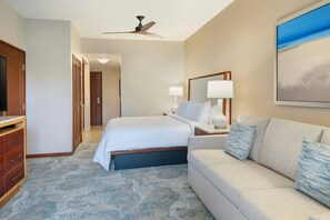 Una habitación de hotel con cama, sofá, televisor, ventilador de techo y un cuadro en la pared.