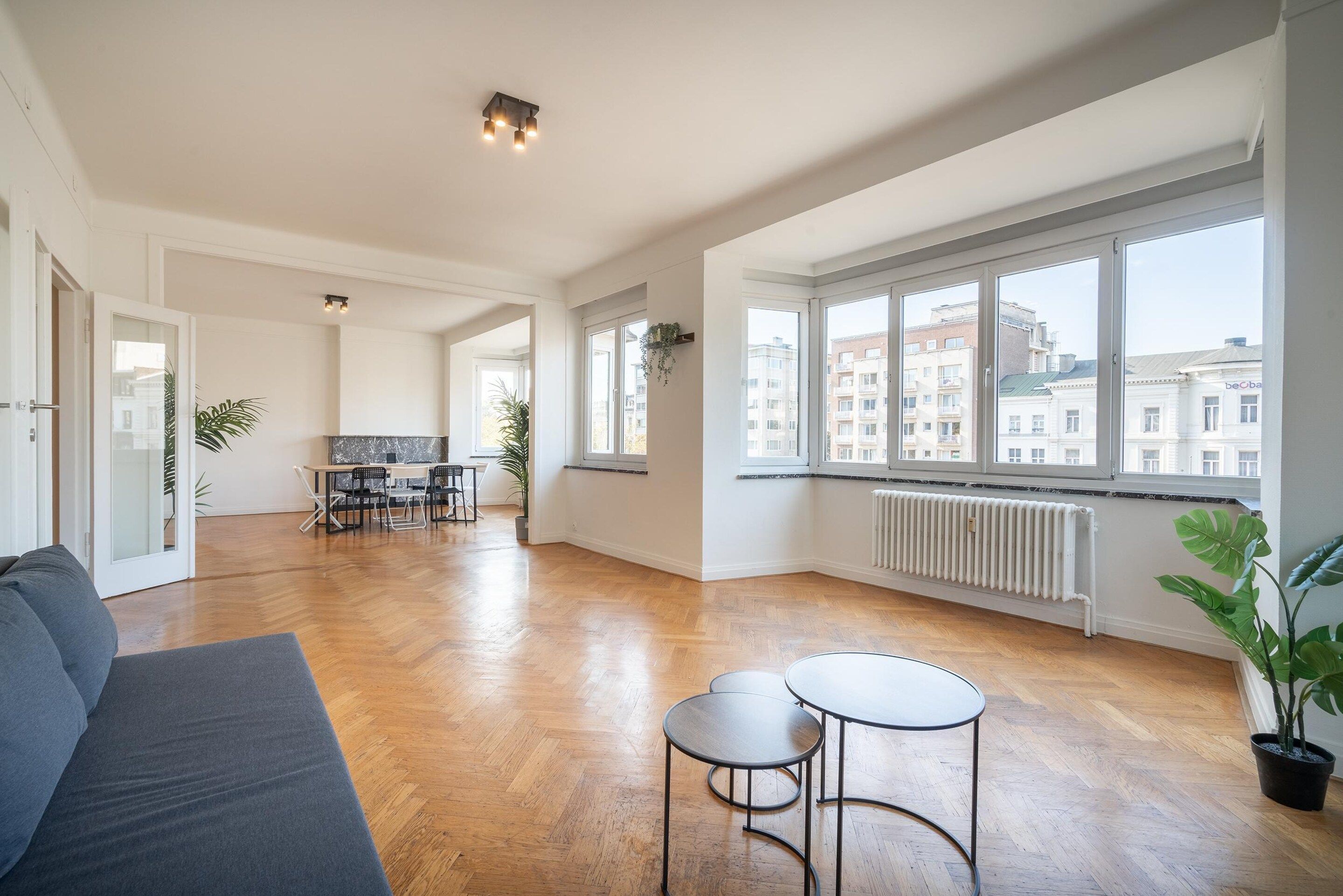 Un salón amplio con suelo de madera, un sofá, una mesa de comedor con sillas y un ventanal con vistas a edificios.