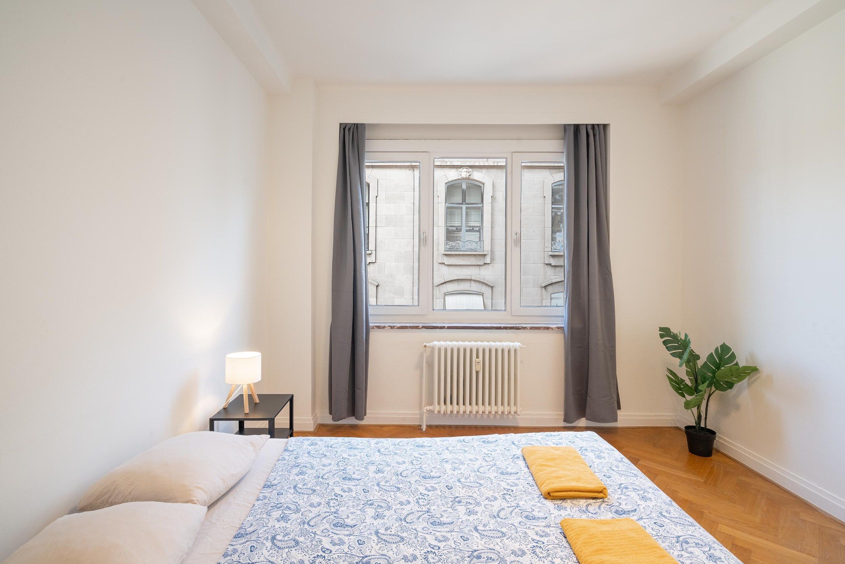 Une chambre à coucher avec un lit, une table de chevet avec une lampe, un radiateur et une fenêtre avec des rideaux.