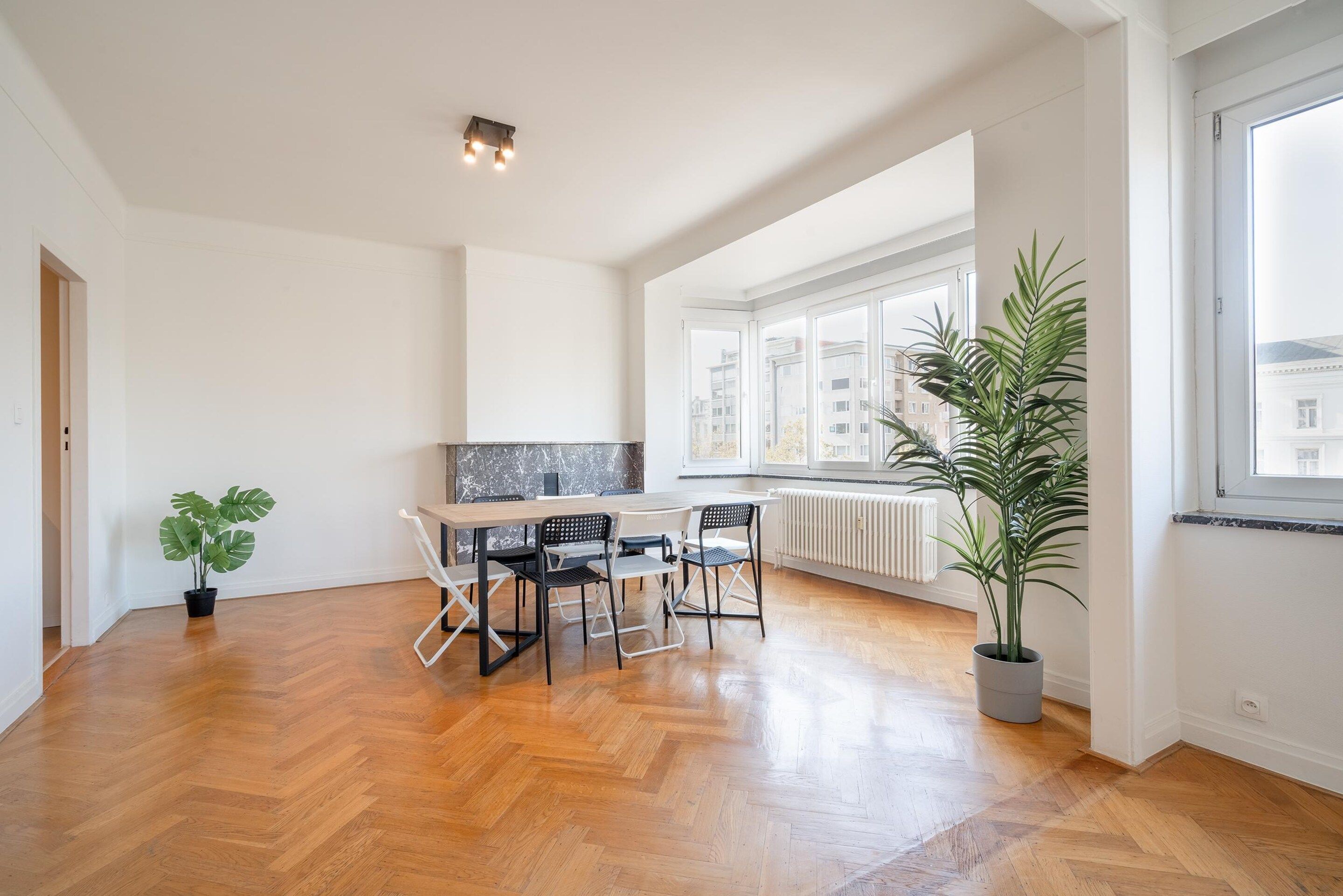 Una habitación amplia con suelo de madera en espiga, una mesa de comedor con sillas, una planta en maceta y un ventanal con vistas a edificios.