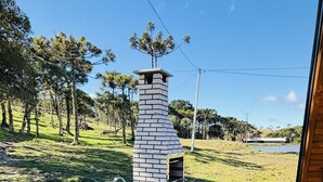 Una cocina exterior de ladrillo con chimenea, una terraza de madera y una zona verde con árboles al fondo.