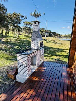 A brick outdoor kitchen with a chimney, a wooden deck, and a grassy area with trees in the background.
