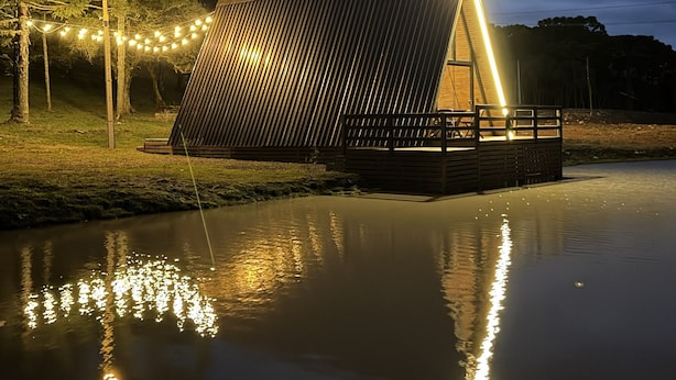 A modern, triangular-shaped building with a flat roof, situated by a calm body of water reflecting lights. - Lake cabin with hot tub and mountain views (Bom Jardim da Serra)