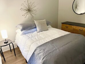 A neatly made bed with a white and gray quilt, a bedside lamp, and a wooden dresser with drawers.